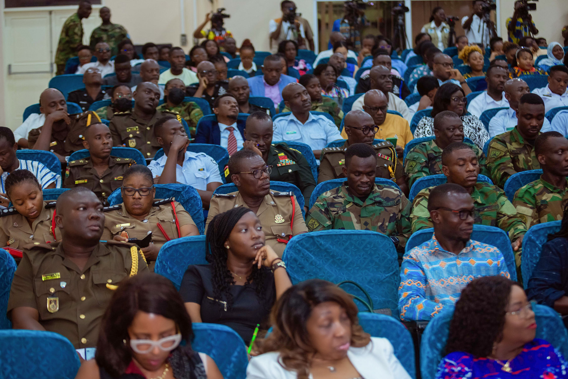 Military, security and media participants seated in the auditorium, with camera operators standing at the back