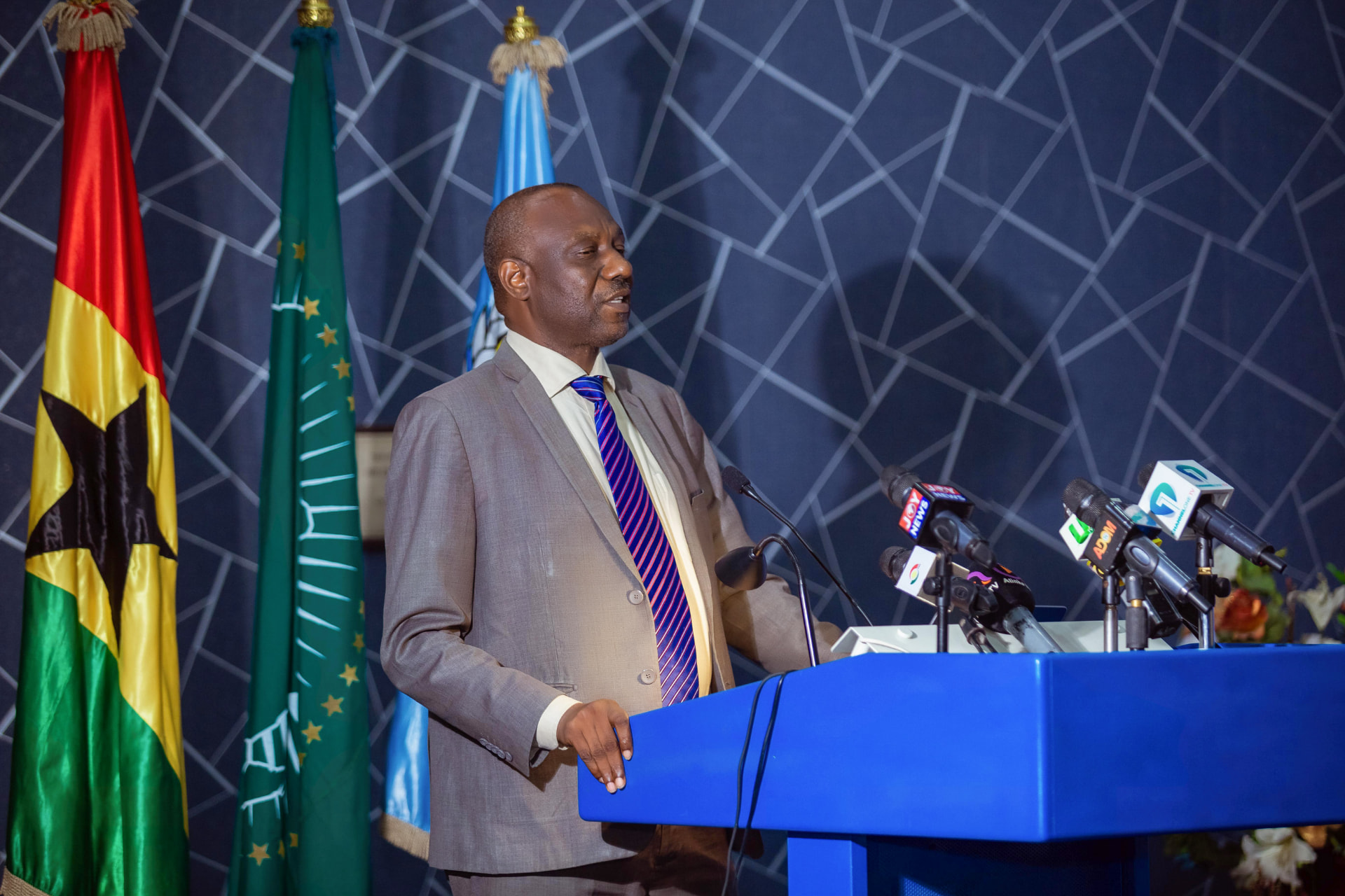 A speaker at the podium with press microphones from Joy News and Adom, beside the flags of Ghana and the African Union